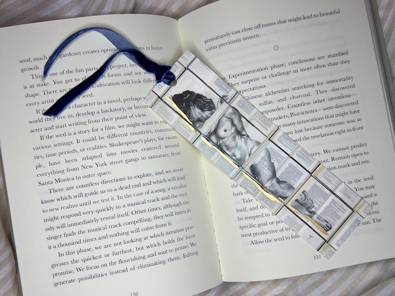 May include: Handmade bookmark with a blue ribbon, featuring a grayscale image of a nude figure. The bookmark is constructed from small, book-page squares, creating a unique, artistic design.