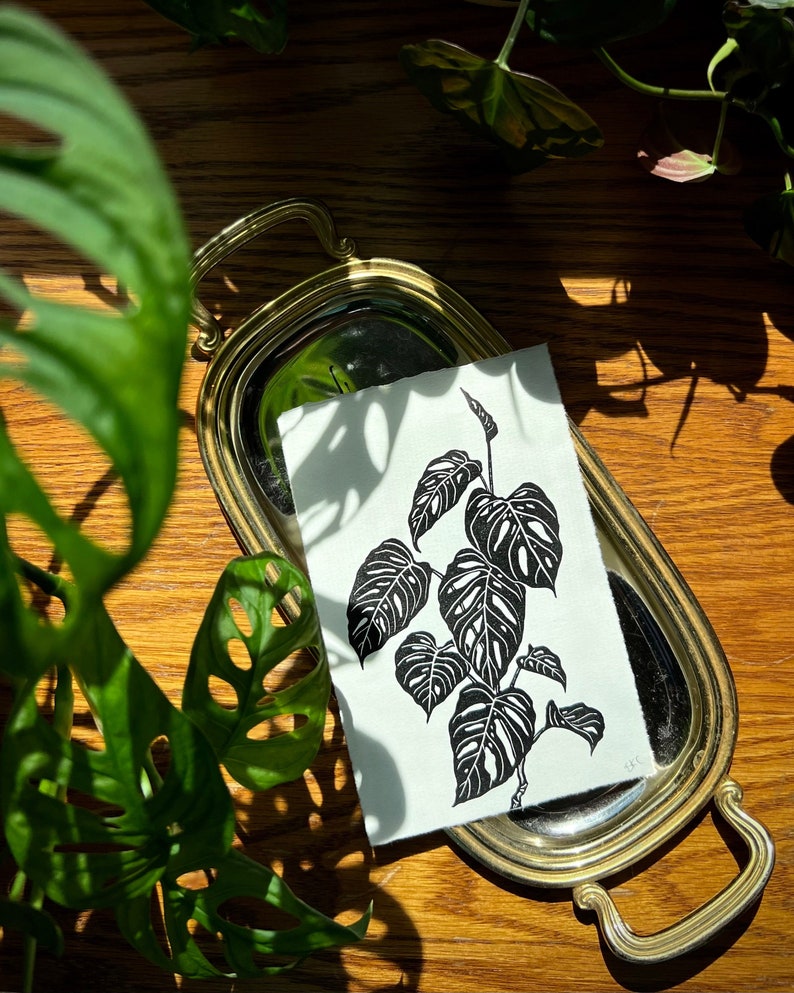 K&ouml;nnte beinhalten: Ein Schwarz-Wei&szlig;-Druck einer Monstera-Pflanze auf wei&szlig;em Hintergrund. Der Druck wird auf einem goldfarbenen Tablett mit Griff pr&auml;sentiert.