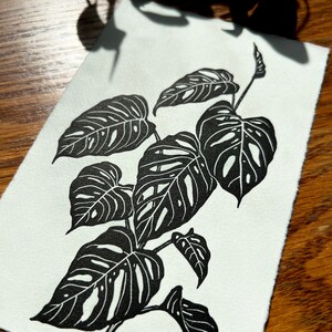 K&ouml;nnte beinhalten: Schwarz-wei&szlig;-Linienkunst-Druck einer Monstera-Pflanze mit gro&szlig;en Bl&auml;ttern. Der Druck ist auf wei&szlig;em Papier.