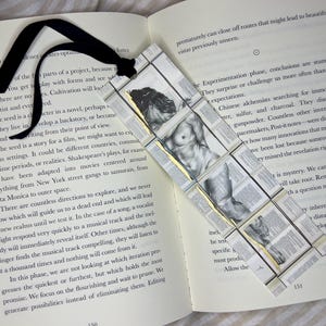 May include: Handmade bookmark featuring a grayscale illustration of a male figure, with a black ribbon. The bookmark is made of small, rectangular pieces with text, and gold accents, placed on an open book.