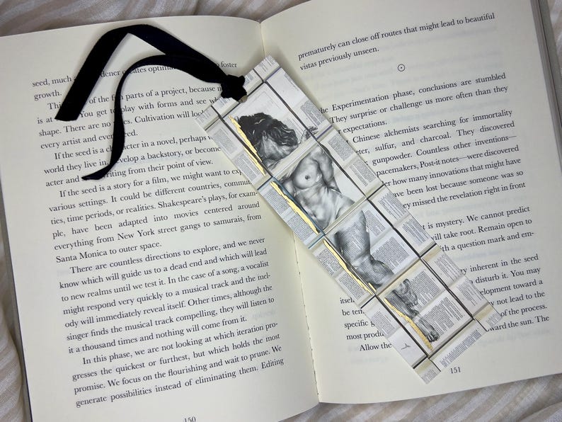 May include: Handmade bookmark featuring a grayscale illustration of a male figure, with a black ribbon. The bookmark is made of small, rectangular pieces with text, and gold accents, placed on an open book.
