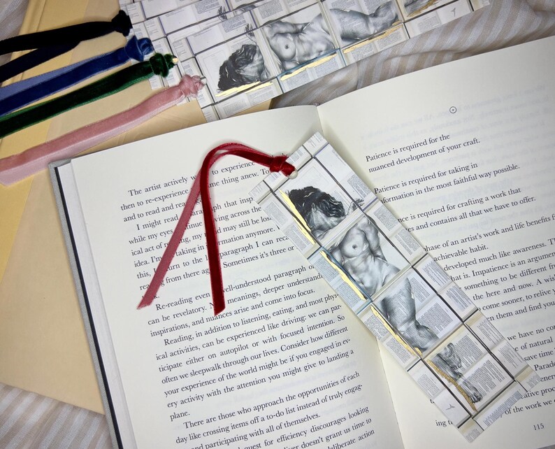 May include: A handmade bookmark featuring a grayscale illustration of a nude figure, set against a backdrop of book pages. The bookmark has a red ribbon and is surrounded by other velvet ribbon bookmarks.