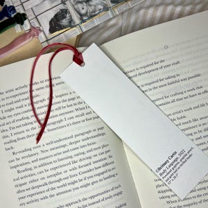 May include: A white rectangular bookmark with a red velvet ribbon attached. The bookmark is placed in an open book. The bookmark has text that reads "Brittany Cartie Body Language, 2022".
