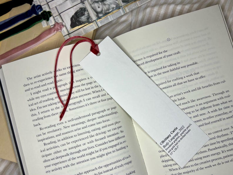 May include: A white rectangular bookmark with a red velvet ribbon attached. The bookmark is placed in an open book. The bookmark has text that reads "Brittany Cartie Body Language, 2022".