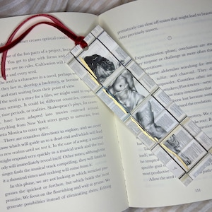 May include: Handmade bookmark featuring a nude figure illustration. The bookmark is constructed from small squares of book pages, with a red ribbon attached. The bookmark is placed in an open book.