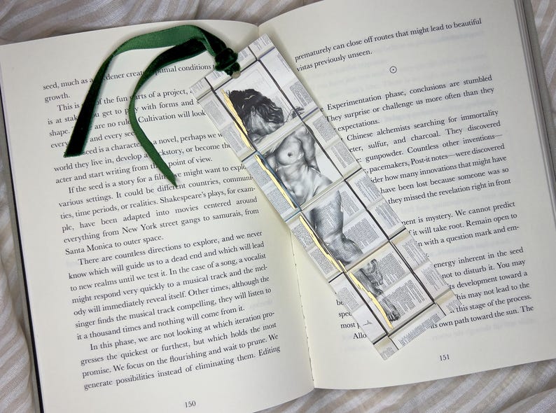 May include: A handmade bookmark featuring a classical nude figure, constructed from small, book-page squares. The bookmark has a green velvet ribbon attached, resting on an open book.