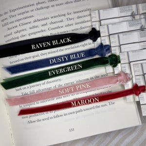 May include: A selection of velvet ribbon bookmarks in various colors: Raven Black, Dusty Blue, Evergreen, Soft Pink, and Maroon. Each bookmark is placed on an open book, with the color name printed in white.