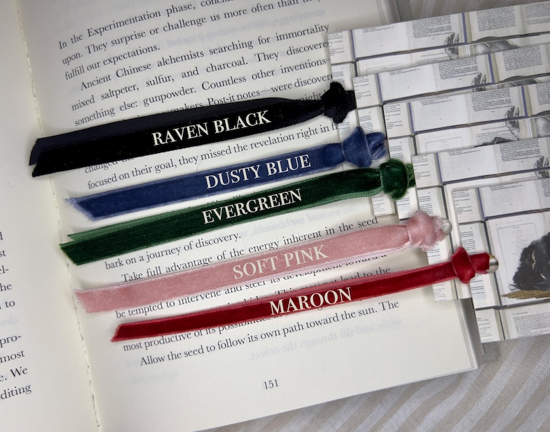 May include: A selection of velvet ribbon bookmarks in various colors: Raven Black, Dusty Blue, Evergreen, Soft Pink, and Maroon. Each bookmark is placed on an open book, with the color name printed in white.