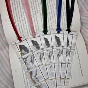 May include: A collection of handmade bookmarks with velvet ribbons in various colors, including red, pink, green, blue, and navy. Each bookmark features a black and white illustration of a classical sculpture. The bookmarks are placed on an open book.