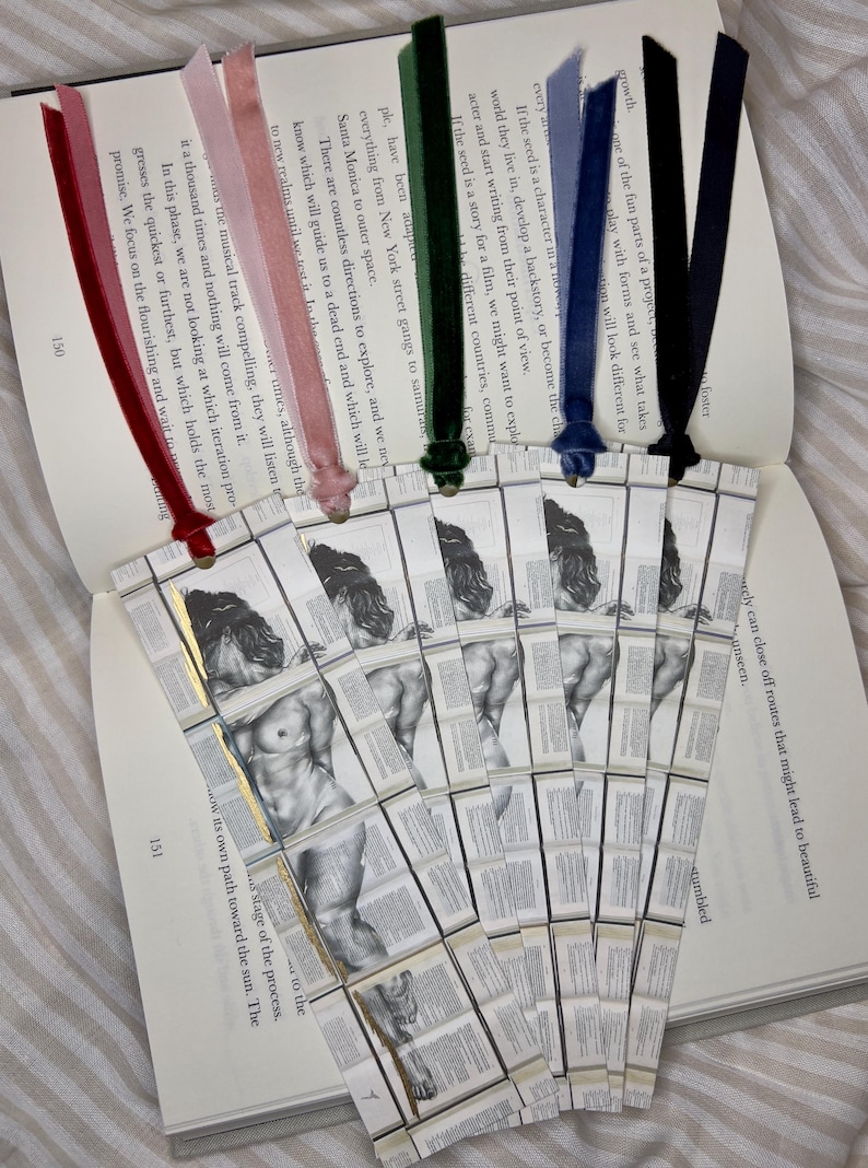 May include: A collection of handmade bookmarks with velvet ribbons in various colors, including red, pink, green, blue, and navy. Each bookmark features a black and white illustration of a classical sculpture. The bookmarks are placed on an open book.