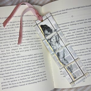 May include: Handmade bookmark featuring a classical nude figure illustration. The bookmark is made of small, rectangular pieces with gold accents and a pink ribbon. Placed on an open book.