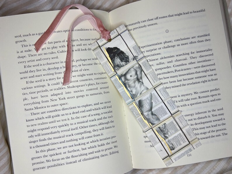 May include: Handmade bookmark featuring a classical nude figure illustration. The bookmark is made of small, rectangular pieces with gold accents and a pink ribbon. Placed on an open book.
