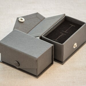 Op de afbeelding: Een grijze kartonnen doos met een scharnierend deksel en een zwart fluwelen interieur. De doos is ontworpen om kleine voorwerpen, zoals sieraden, in te bewaren.