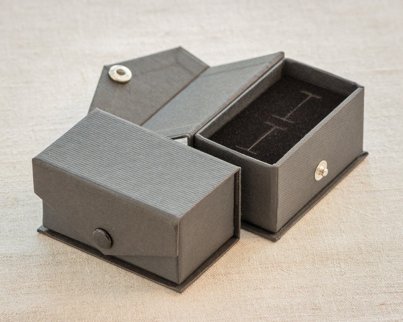 Op de afbeelding: Een grijze kartonnen doos met een scharnierend deksel en een zwart fluwelen interieur. De doos is ontworpen om kleine voorwerpen, zoals sieraden, in te bewaren.