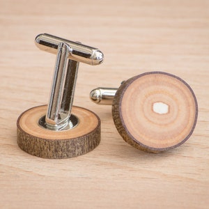 Op de afbeelding: Een paar zilveren manchetknopen met houten bases. Het hout is lichtbruin van kleur met een donkerbruine ring rond de rand. De manchetknopen liggen op een houten oppervlak.