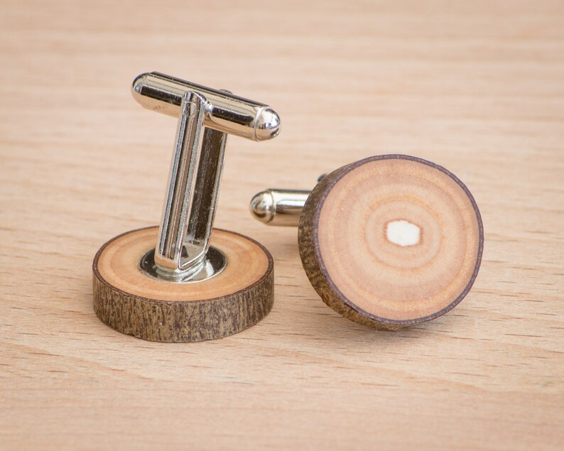 Op de afbeelding: Een paar zilveren manchetknopen met houten bases. Het hout is lichtbruin van kleur met een donkerbruine ring rond de rand. De manchetknopen liggen op een houten oppervlak.