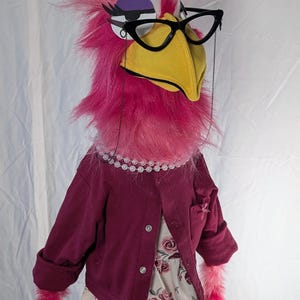 May include: A whimsical bird puppet with pink and white feathered head, wearing black glasses, a pearl necklace, and a burgundy jacket over a floral dress. The puppet has a yellow beak and pink feathered arms.