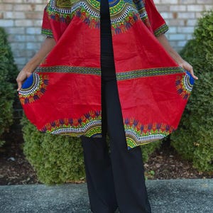 May include: A vibrant red open-front dashiki-style jacket with short sleeves. The jacket features a colorful patterned design in blue, green, yellow, and white along the edges and collar. The model is wearing black pants and sandals.