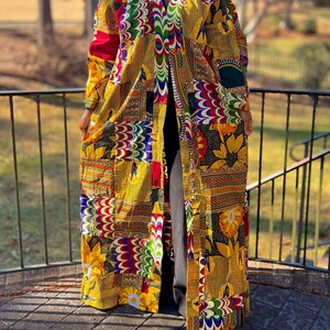 May include: A long, patchwork-style duster coat in vibrant colors. The coat features a mix of patterns, including floral, geometric, and abstract designs in yellow, red, green, and blue. The coat is open in the front and has long sleeves.