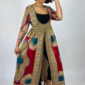Op de afbeelding: Een lange, open jurk met een levendige Afrikaanse print in rood, geel, groen en goud. De jurk heeft een vierkante halslijn, driekwartmouwen en een getailleerde taille. Het kledingstuk wordt gedragen over een zwarte top en broek.