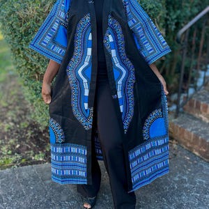May include: A long, open-front black kimono with vibrant blue and white geometric patterns. The kimono features short sleeves and is worn over black pants and a black top. The design includes intricate details and a bold color contrast.