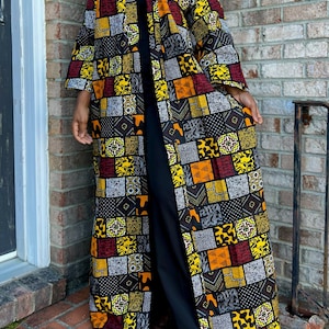 May include: A long, open-front kimono-style robe with a vibrant patchwork design. The robe features a mix of geometric and abstract patterns in black, yellow, orange, and red. The robe is worn over a black outfit.