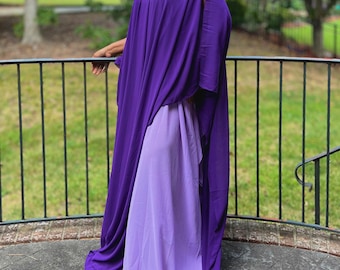 May include: A flowing, floor-length dress in shades of purple. The outer layer is a deep violet, with a cape-like design and arm slits. Underneath, a lighter lavender dress peeks through, creating a layered effect. The dress is made of a soft, draping fabric.