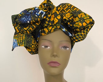 May include: A vibrant head wrap featuring a bold pattern in yellow, blue, green, and black. The fabric is wrapped and tied to create a voluminous, decorative style. The head wrap is displayed on a mannequin head.