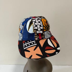 May include: A colorful baseball cap with a patchwork design featuring blue, white, orange, red, and brown geometric patterns. The brim of the cap is black with a peach and white geometric pattern.