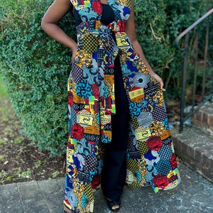 May include: A sleeveless, floor-length garment with a vibrant patchwork design in red, blue, yellow, and black. The garment features a V-neck and a matching belt. The person is wearing black pants and sandals.