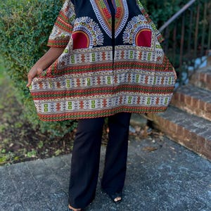 May include: A colorful, patterned dashiki-style jacket with short sleeves. The jacket features a black base with intricate designs in red, green, and white. The person is wearing black pants and black sandals.
