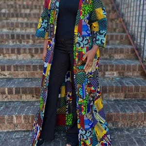 Puede incluir: Un abrigo largo abierto por delante con un vibrante diseño de patchwork de varios patrones geométricos y abstractos en tonos brillantes de rojo, azul, amarillo y verde. La prenda tiene mangas tres cuartos y un dobladillo recto hasta el suelo.
