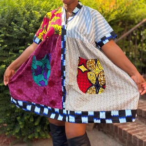 May include: A short-sleeved dress featuring a patchwork design with a mix of fabrics. One side displays a deep magenta floral pattern, while the other side features light blue and white stripes. It includes two large pockets and a blue and black checkered hem.