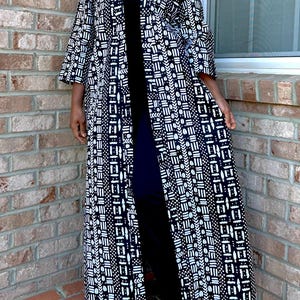 May include: A long, open-front black and white patterned kimono-style robe. The robe features a geometric design and three-quarter sleeves. The person is wearing a black outfit underneath. The background is a brick wall and a window.