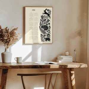 May include: A framed 2026 calendar with a cream background and black illustrations of forest animals and foliage. The calendar is displayed on a wooden desk with a vase of dried plants, a mug, and books. The wall is painted a neutral color.