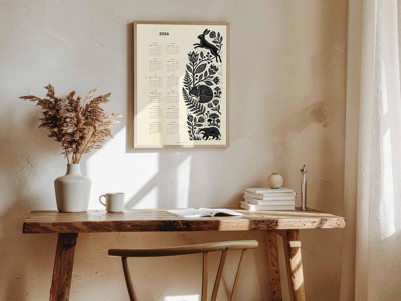 May include: A framed 2026 calendar with a cream background and black illustrations of forest animals and foliage. The calendar is displayed on a wooden desk with a vase of dried plants, a mug, and books. The wall is painted a neutral color.