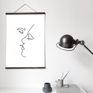 May include: A minimalist art print featuring a black line drawing of two faces in profile, appearing to kiss. The artwork is displayed on a white background and held by a dark brown wooden frame. The print hangs on a white wall.