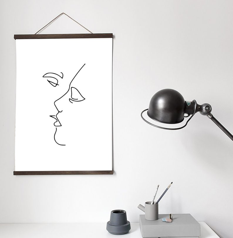 May include: A minimalist art print featuring a black line drawing of two faces in profile, appearing to kiss. The artwork is displayed on a white background and held by a dark brown wooden frame. The print hangs on a white wall.