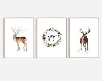 May include: Set of three framed watercolor prints featuring winter themes. One print shows a deer with large antlers, another a wreath with the word "joy", and the third a deer with large antlers. The prints have a white background.