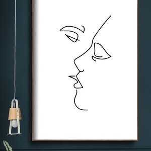 May include: A framed minimalist art print featuring a black line drawing of two faces in profile, appearing to be kissing. The artwork is set against a white background, with a simple, elegant aesthetic. The frame is a dark brown color.