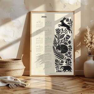 May include: A framed 2026 calendar print with a cream background. The calendar features a black and white illustration of forest animals and foliage. A jumping rabbit, sleeping fox, and badger are depicted.