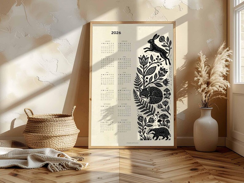 May include: A framed 2026 calendar print with a cream background. The calendar features a black and white illustration of forest animals and foliage. A jumping rabbit, sleeping fox, and badger are depicted.