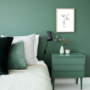 May include: A bedroom scene with a green wall, bed, and nightstand. The bed has white and green pillows. A framed line drawing of a face hangs on the wall. A small lamp and vase with flowers sit on the nightstand.