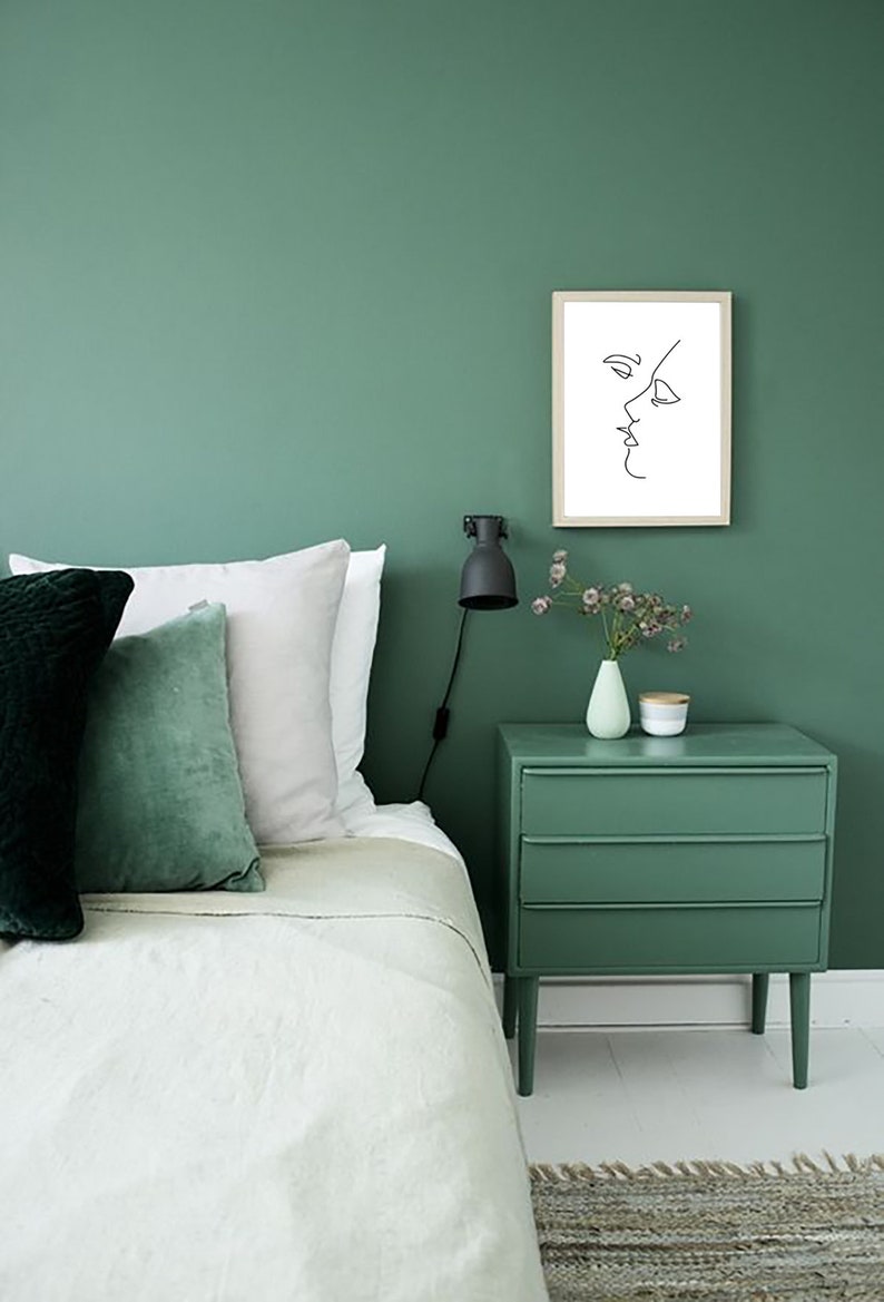 May include: A bedroom scene with a green wall, bed, and nightstand. The bed has white and green pillows. A framed line drawing of a face hangs on the wall. A small lamp and vase with flowers sit on the nightstand.