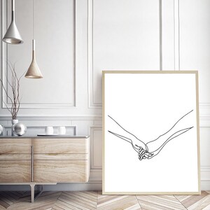Może przedstawiać: Oprawiony wydruk artystyczny z minimalistycznym rysunkiem liniowym dw&oacute;ch trzymających się dłoni. Grafika jest czarna na białym tle, z jasnobrązową ramą. Wydruk jest eksponowany w pokoju z drewnianą szafką.