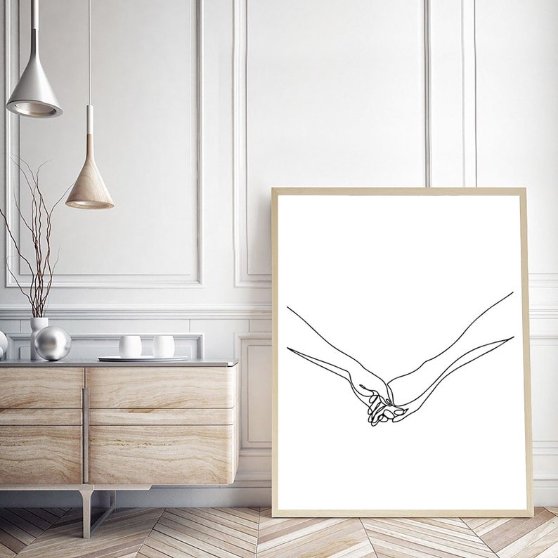 Może przedstawiać: Oprawiony wydruk artystyczny z minimalistycznym rysunkiem liniowym dw&oacute;ch trzymających się dłoni. Grafika jest czarna na białym tle, z jasnobrązową ramą. Wydruk jest eksponowany w pokoju z drewnianą szafką.