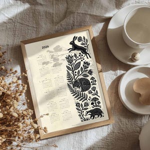 May include: A framed 2026 calendar with a nature-themed design. The black and white illustration features a leaping rabbit, sleeping fox, and badger amidst floral and botanical elements. A cup and saucer are on the right.