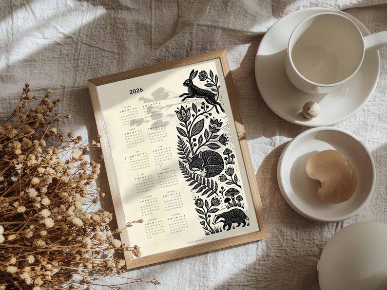 May include: A framed 2026 calendar with a nature-themed design. The black and white illustration features a leaping rabbit, sleeping fox, and badger amidst floral and botanical elements. A cup and saucer are on the right.
