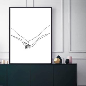 Może przedstawiać: Czarny obraz w ramie przedstawiający minimalistyczny rysunek liniowy dw&oacute;ch trzymających się dłoni. Grafika to proste, ale eleganckie przedstawienie połączenia i wsparcia. Dłonie narysowane są ciągłą linią na białym tle.