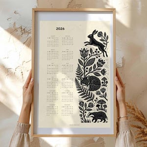 May include: A framed 2026 calendar print with a cream background. The calendar features a black and white illustration of a rabbit, fox, and badger among floral and botanical elements. The frame is a light wood color.